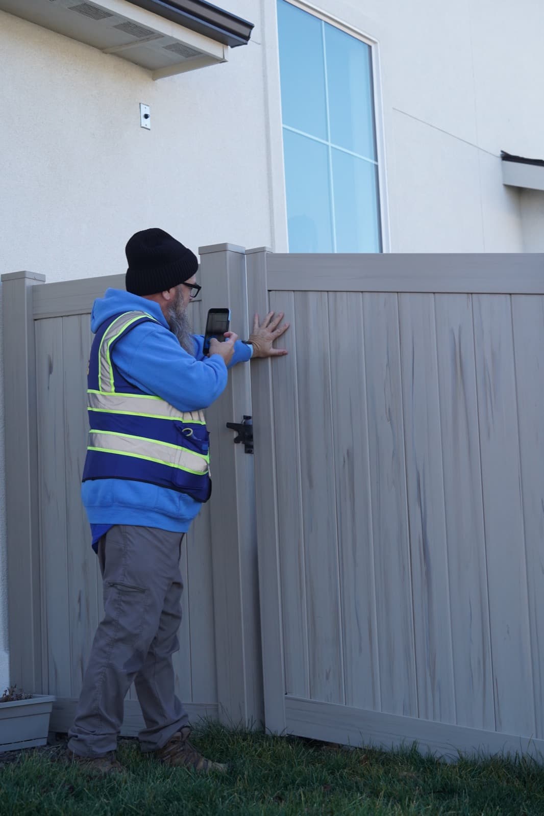A Lawn Dogs technician photographing a closed side gate with one hand pressed flat against it to confirm the latch is secure