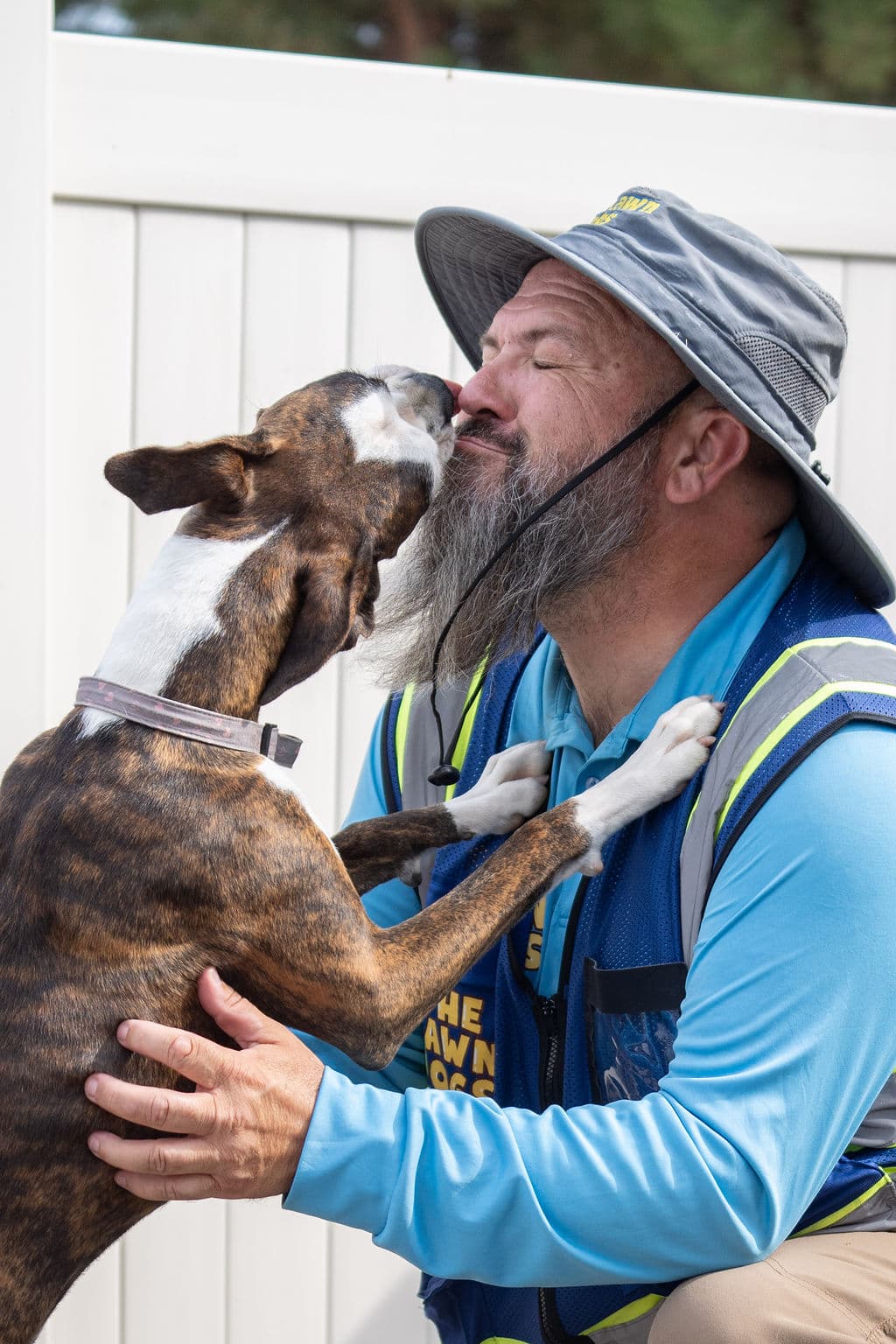 Donovan in a branded Lawn Dogs vest crouching in a yard while a brindle boxer puts its paws on his chest and licks his face