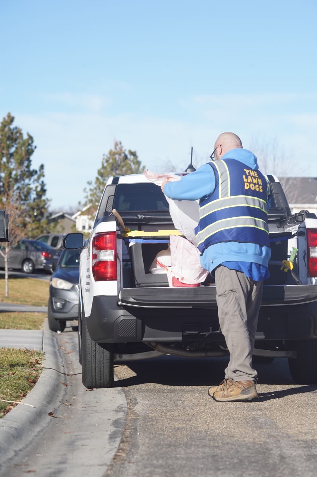 A Lawn Dogs technician in a branded vest loading bagged waste into the bed of a company truck on a residential street
