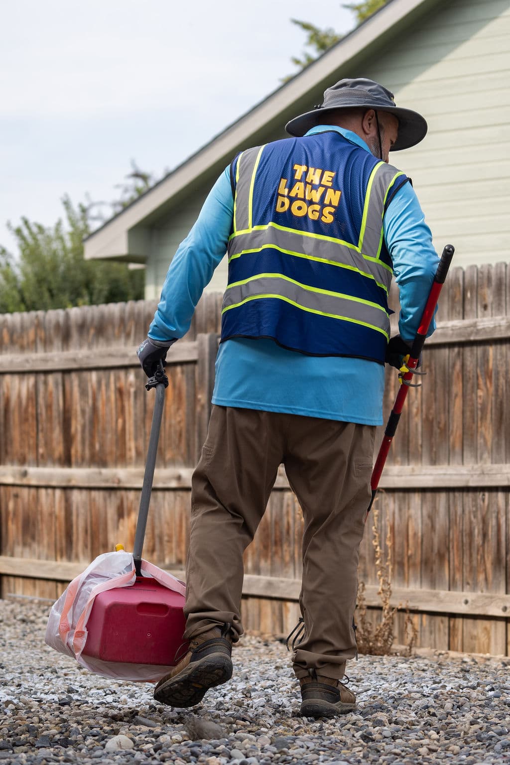 A Lawn Dogs technician in a branded hi-vis vest carrying bagged waste and scooping tools out of a fenced back yard
